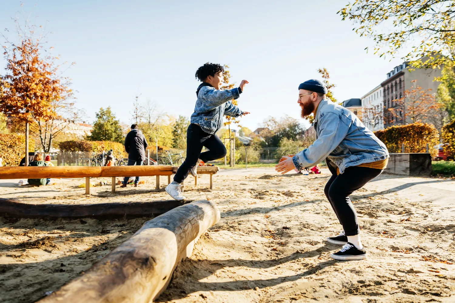 A young boy leaping into his fathers arms from a log while messing around in a playground at the park together.